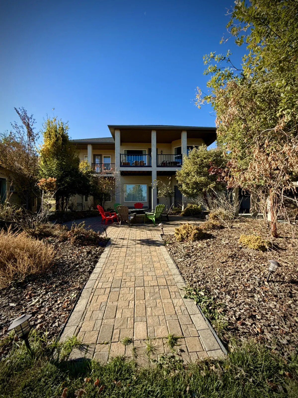 A landscaped yard with a prominent firepit encircled by red and green chairs, set near a pathway and a welcoming house. The area invites both group gatherings and quiet moments, enhanced by natural sunlight and greenery.