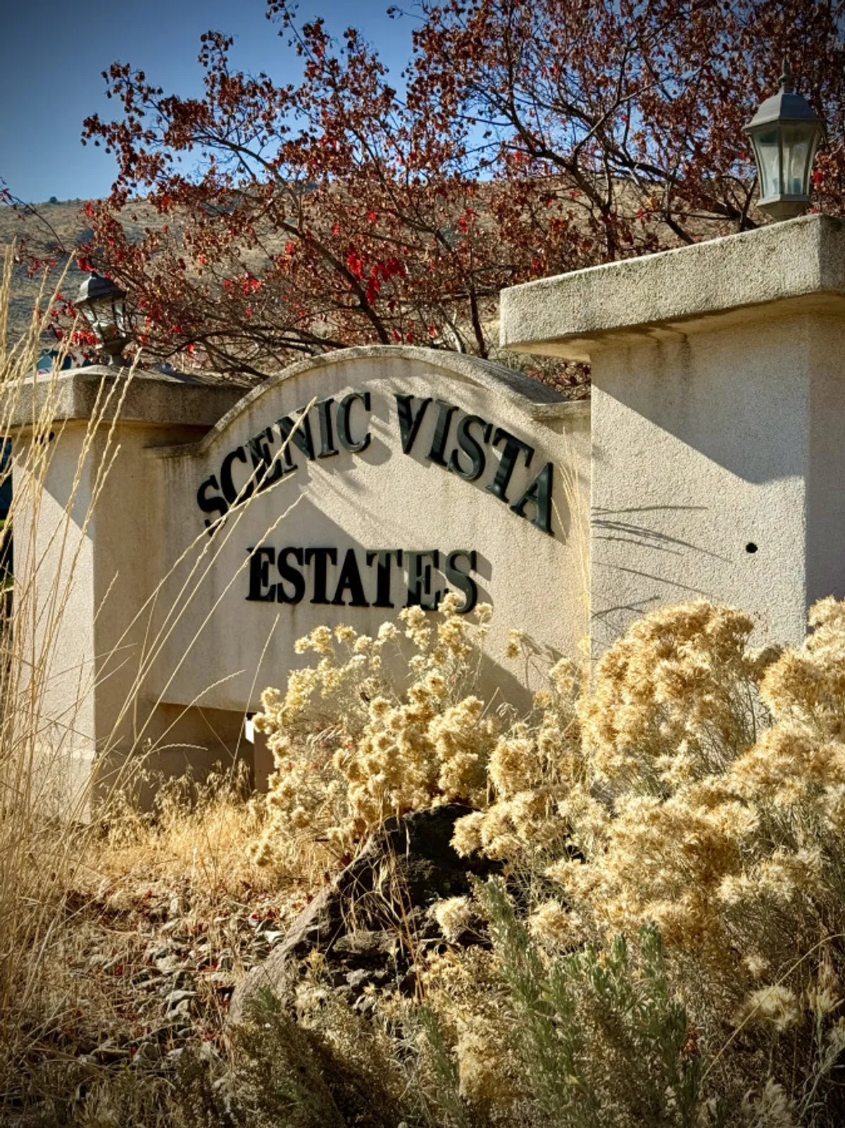 A tranquil estate entrance sign surrounded by lush foliage and natural landscape features, with softly rolling hills in the background. The environment suggests access to scenic trails, wildflowers, and outdoor exploration in warmer months.