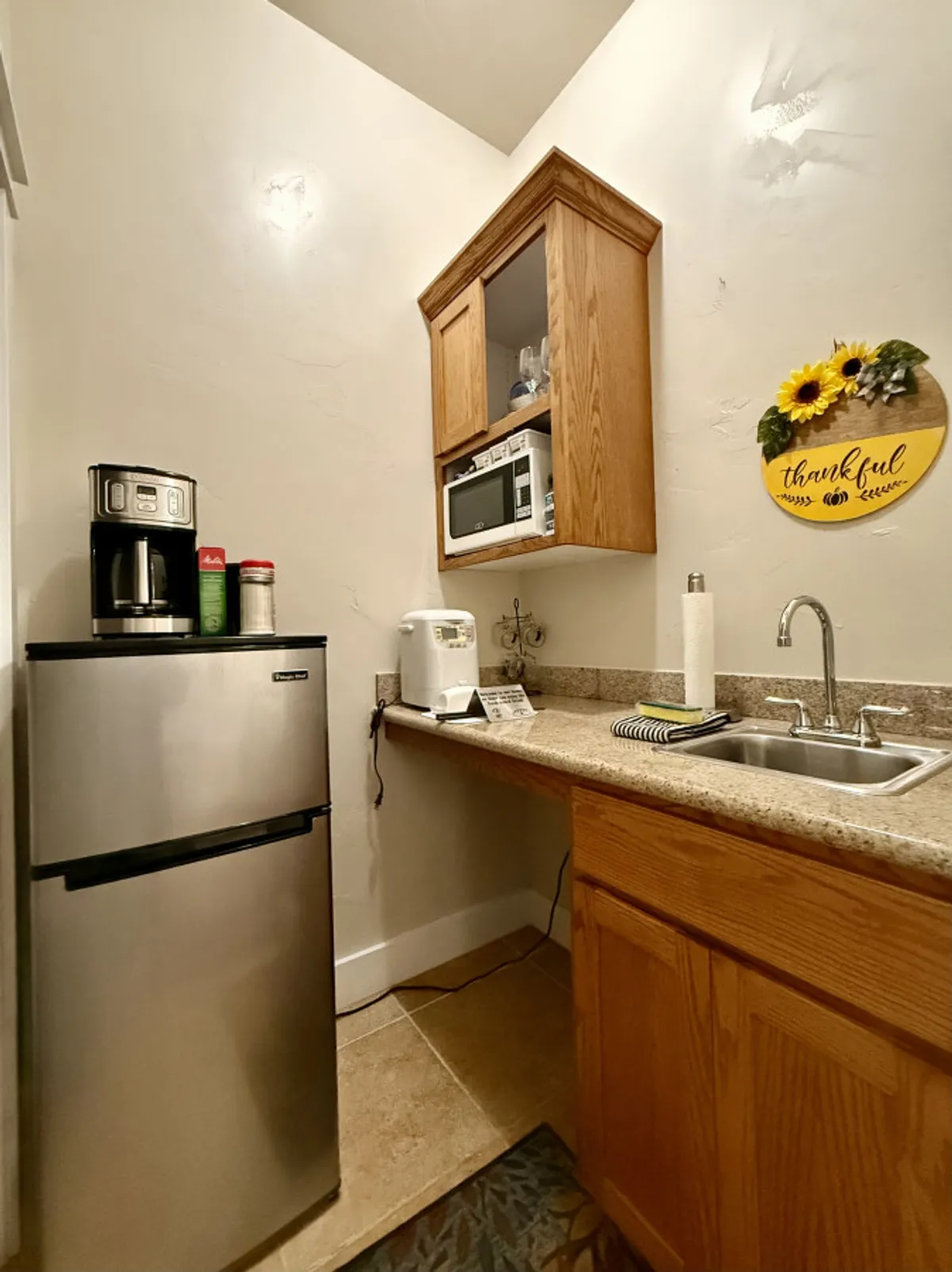 A bright kitchenette area featuring a coffee maker, microwave, sink, and cheerful sunflower wall decor. The counters are uncluttered and the space looks ready for preparing coffee, snacks, or light meals. The pleasant lighting adds an inviting feel, suggesting morning rituals or casual meal prep.
