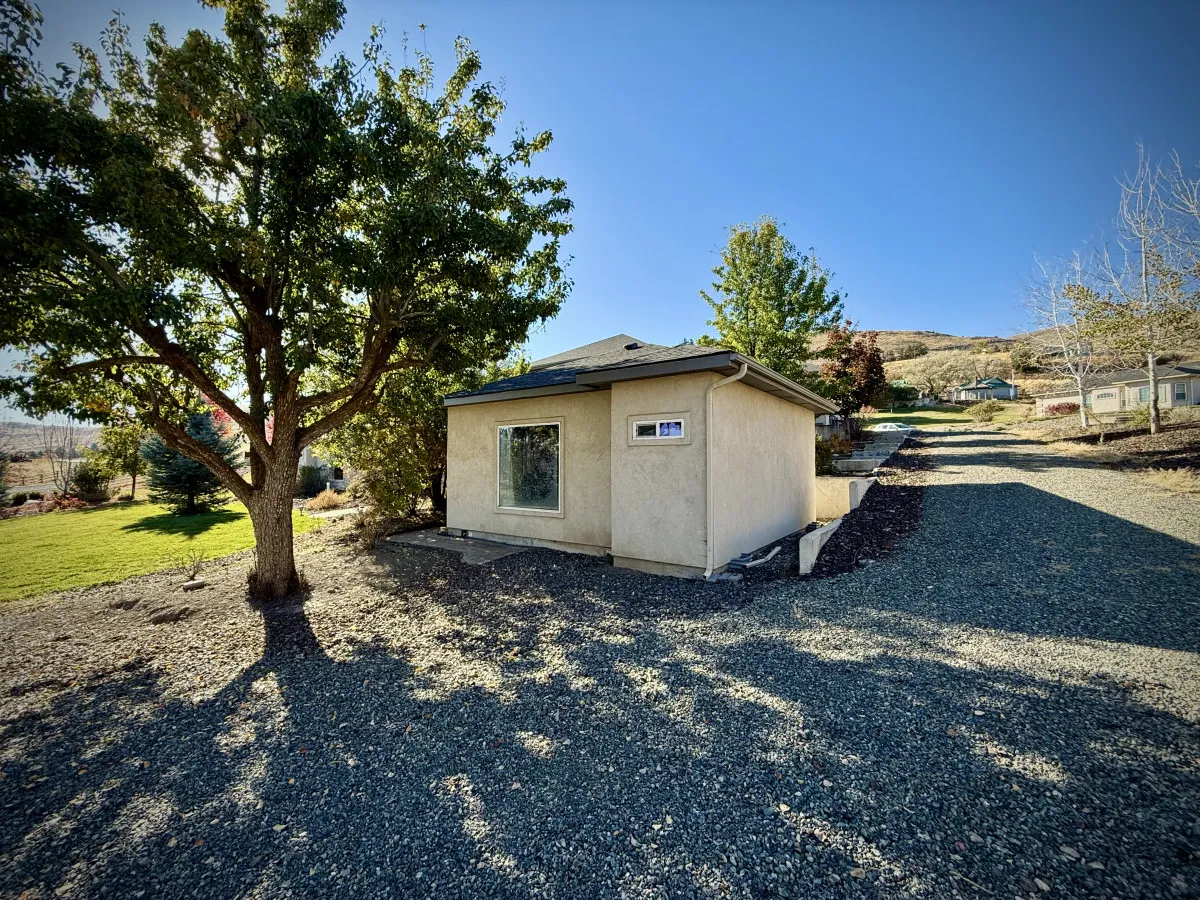 A peaceful exterior scene featuring a small house nestled beside a large tree, with a gravel pathway leading up to it. The image suggests a tranquil spot for relaxing under the tree or hosting a small gathering, with children playing in the yard and sunsets visible from the porch.