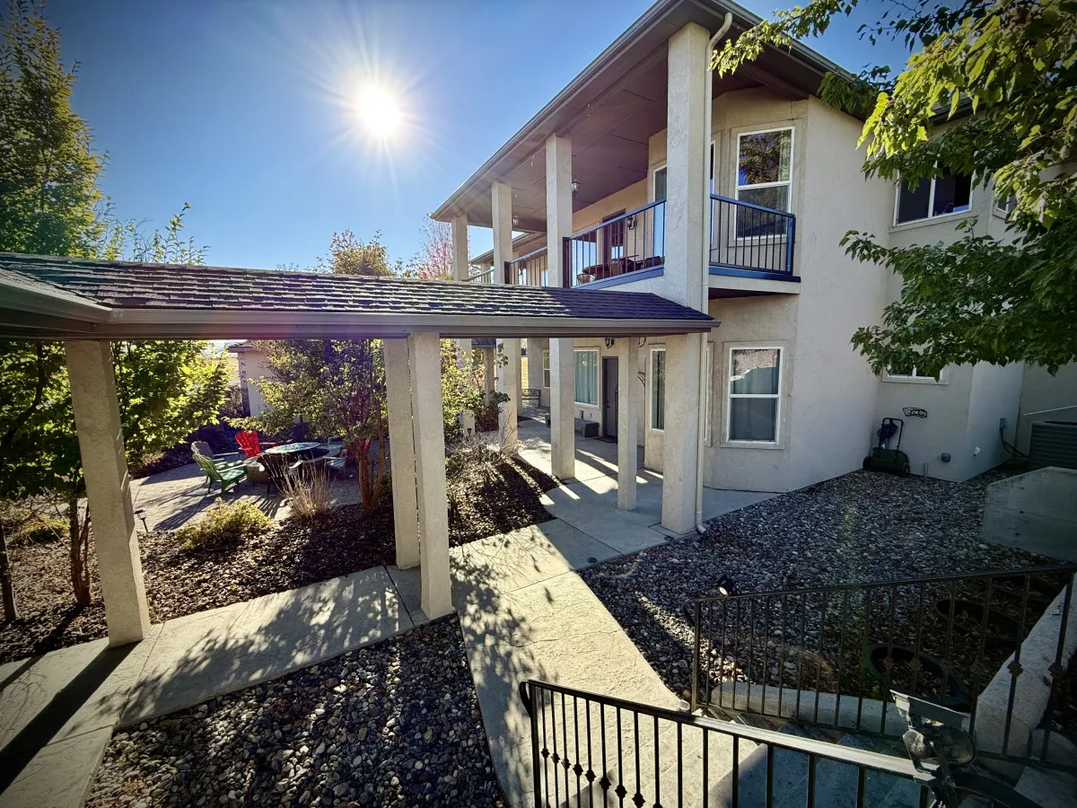 The photo highlights sunlight streaming through trees onto a welcoming front porch with outdoor seating. The porch area is perfect for enjoying morning coffee or evening conversations, emphasizing the connection between indoor comfort and the outdoors.