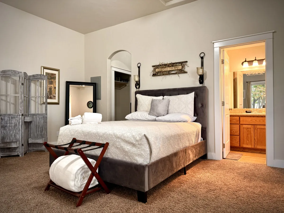 The image showcases a sunlit studio with a plush queen bed dressed in soft linens, a stylish dressing mirror that reflects the inviting space, and a peek into the fresh, modern bathroom. Natural light pours through large windows, creating a warm, serene atmosphere perfect for relaxation or unwinding with a good book.
