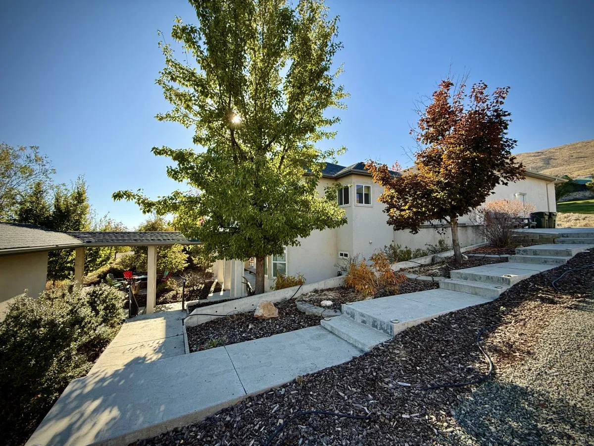 An inviting scene showing a green, landscaped pathway with steps leading up to the house, shaded by mature trees. The garden path and patio area are perfect for strolls, outdoor play, and enjoying fresh air together.