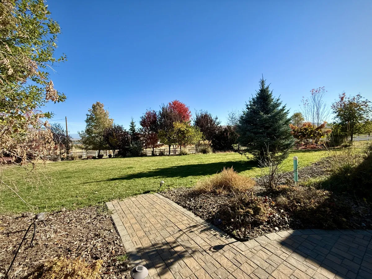 A serene view of a green lawn surrounded by flowering shrubs, evergreen trees, and a curving pathway. The inviting landscape suggests space for outdoor gatherings, games, and peaceful relaxation within nature.