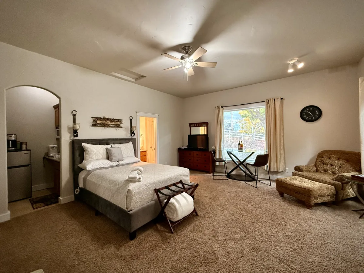 The photo captures a cozy studio corner featuring a queen bed, a comfortable armchair, a dining table, and a broad window offering a view of the outdoors. The space feels intimate and perfect for relaxation or unwinding with a book.