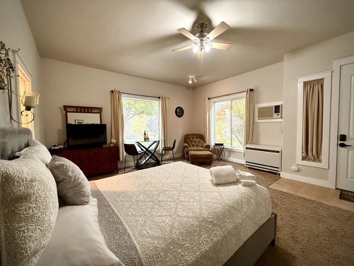 This image presents a studio layout featuring a neatly made bed with decorative linens, an armchair, a small dining table with chairs, and large windows that bathe the room in natural light. The windows offer pleasing views of the landscaped yard outside, enhancing the sense of tranquility.