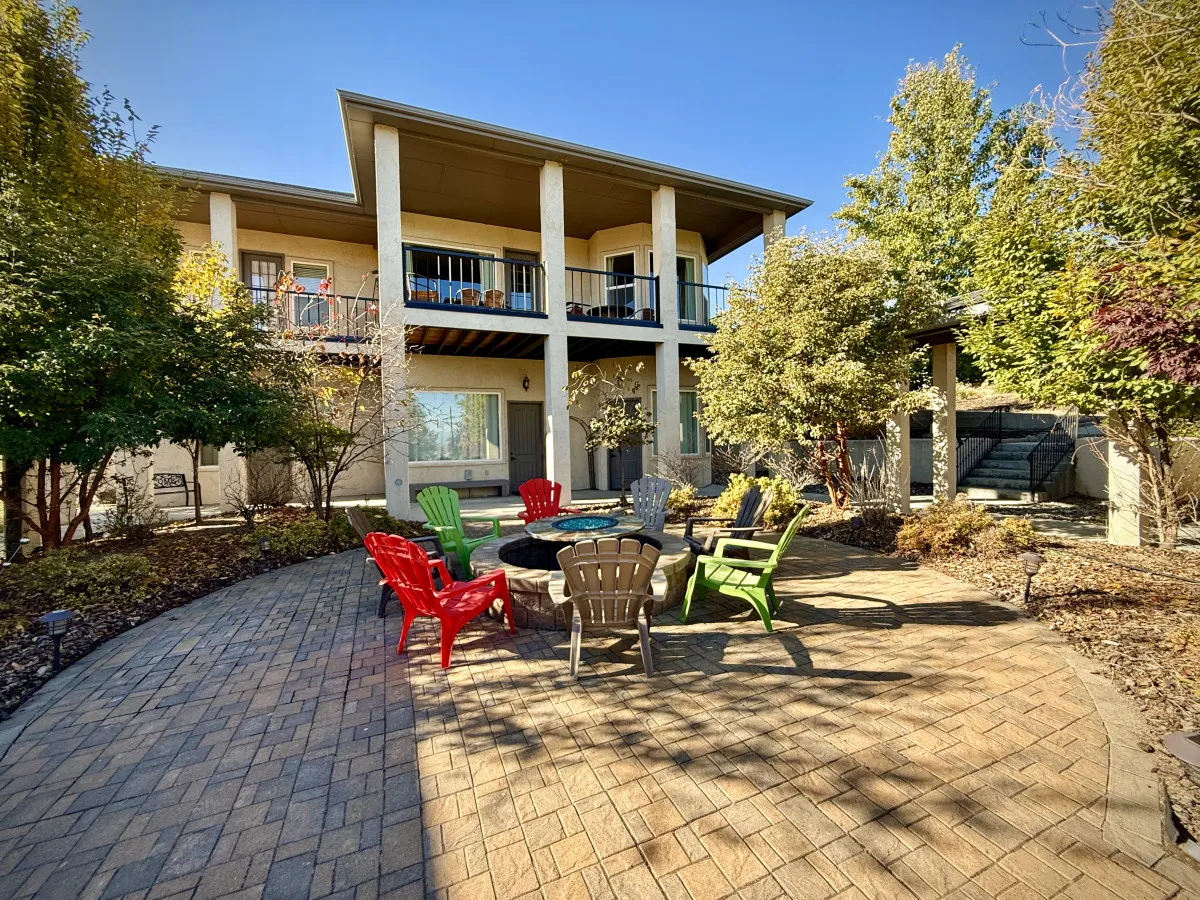 A shared outdoor space featuring a fire pit surrounded by colorful chairs, set amidst lush greenery and a two-story building in the background. The space is clearly designed for gatherings and relaxation, with ample seating and inviting landscaping.
